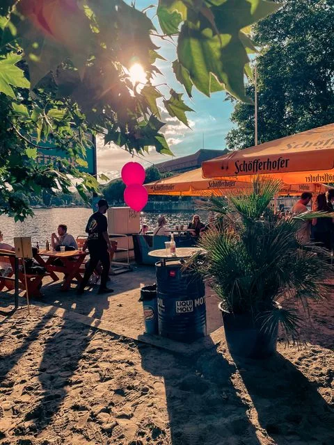 Sonniger Außenbereich des Gestrandet an der Spree mit orangenen Sonnenschirmen, Sitzplätzen im Sand und Gästen im Schatten.